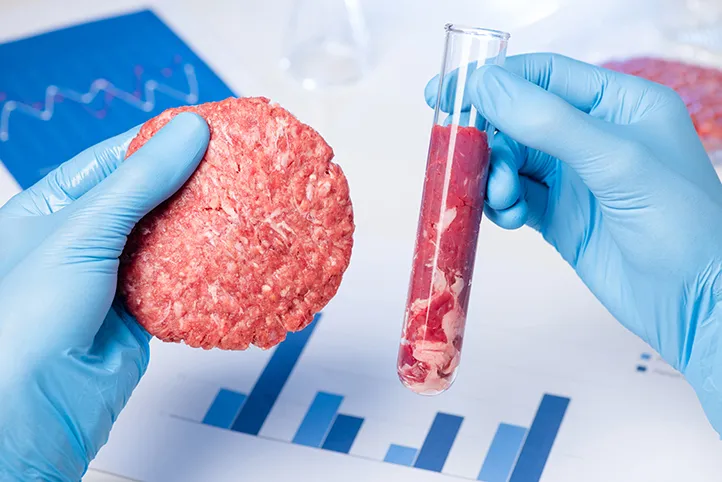 Two gloved hands holding a burger patty and a test tube full of meat